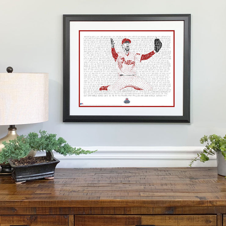Matted and framed wall word art print of Brad Lidge celebrating last out in 2008 World Series hangs on wall over wood table.