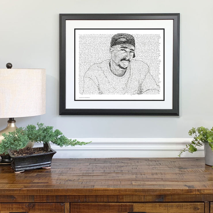 Matted and framed word art drawing of Tupac Shakur in t-shirt and cap, handwritten with selected rap lyrics, hangs on wall.
