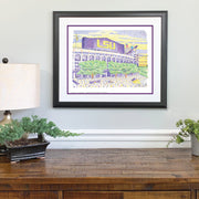 Framed LSU Tiger drawing of stadium in yellow and purple with green trees made of words on wall above wood desk. 