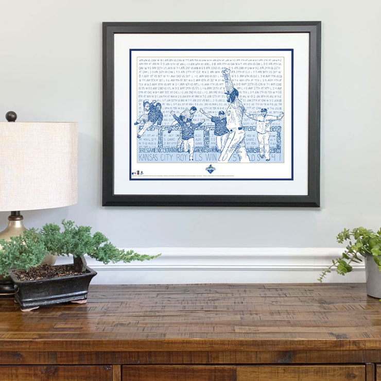 Matted and framed wall word art print depicting 2015 Kansas City Royals World Series win hangs on wall over wooden table.