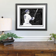 Matted and framed word art Aaron Judge poster hangs on wall above wooden table with small lamp and plants.