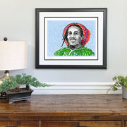 Matted and framed word art portrait of Bob Marley hangs over wooden table, one of the best Bob Marley gifts.
