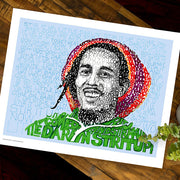 Unframed print of Bob Marley artwork is portrait of him formed by handwritten “Stir It Up” lyrics, lying flat on wood table.