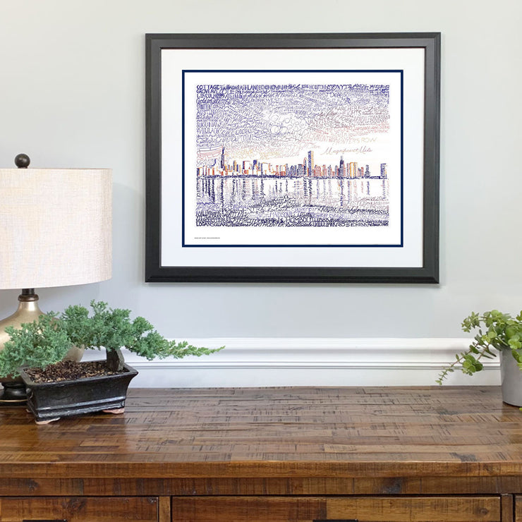 Matted and framed Chicago wall art print, the skyline handwritten with local places and landmarks, hangs on wall above table.