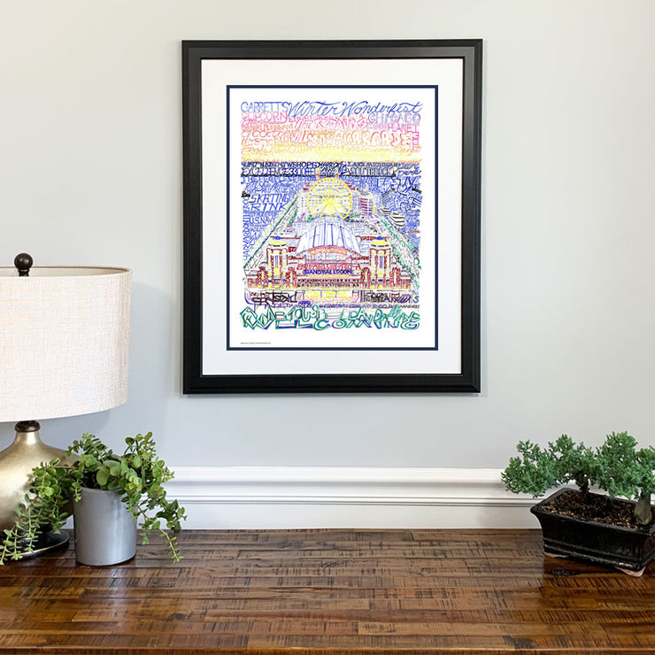 Matted and framed print of Navy Pier art, handwritten with pier history and names of attractions, hangs on wall above table.