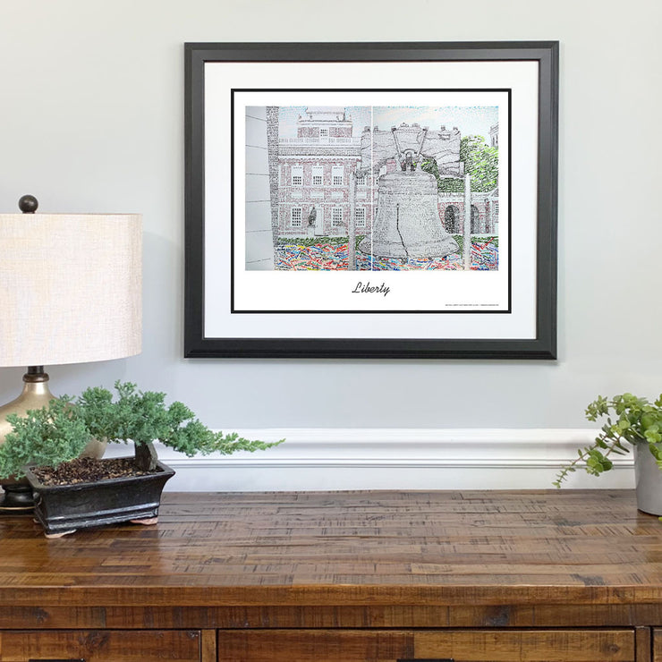 Matted and framed word art scene of Liberty Bell in front of Independence Hall hangs on wall over wooden table.