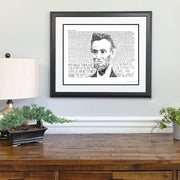 Matted and framed print of Abraham Lincoln art hangs on wall above wooden table with small lamp and plants.